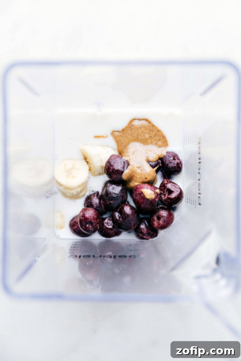 All the essential ingredients for a healthy cherry smoothie in the blender, including almond milk, frozen cherries, almond butter, and frozen bananas, perfectly arranged and ready to be blended together for a nutritious and delicious beverage.