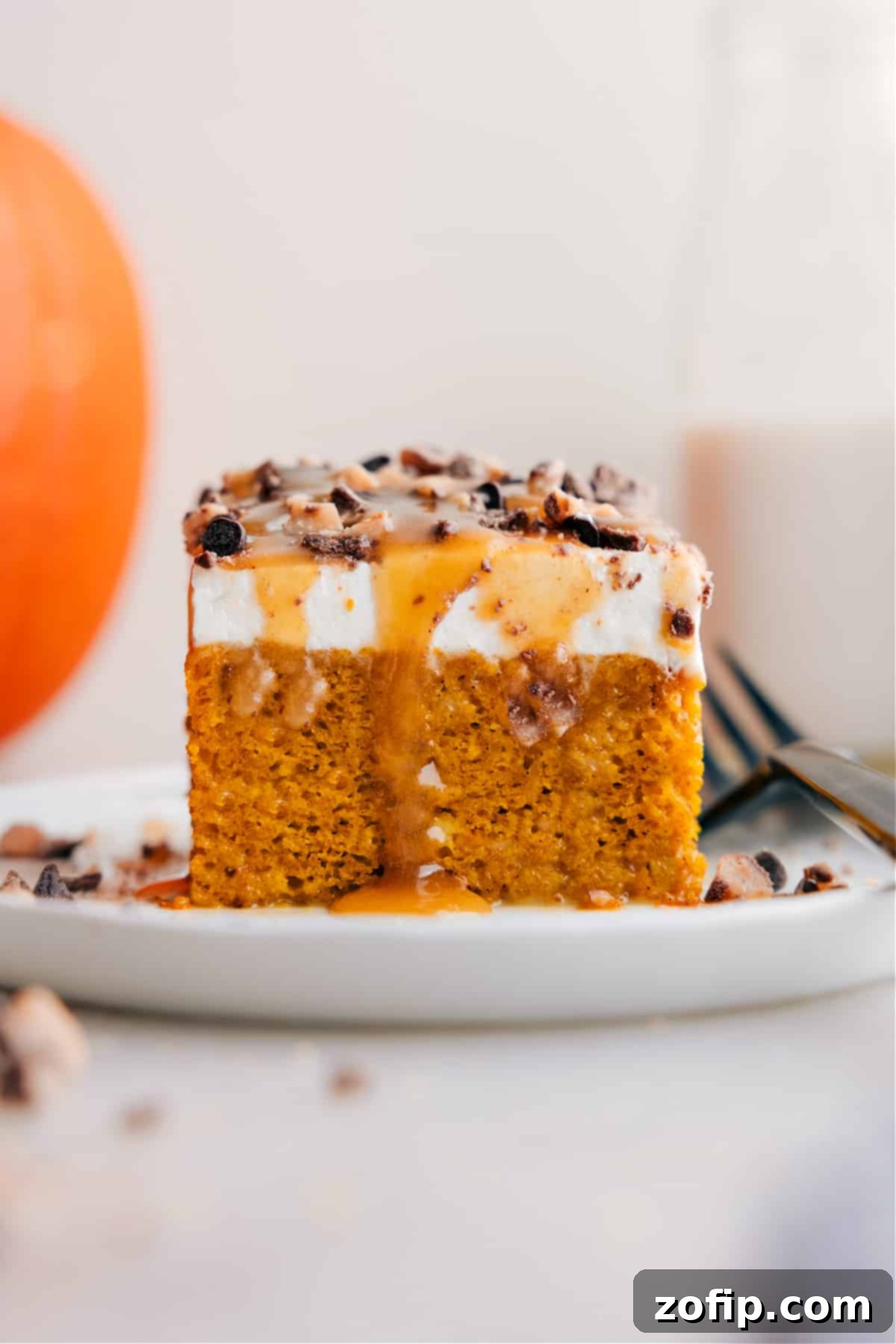The Best Pumpkin Poke Cake Recipe for Fall A perfectly cut slice of Pumpkin Poke Cake, revealing its moist interior soaked with caramel, topped with whipped cream and toffee bits.
