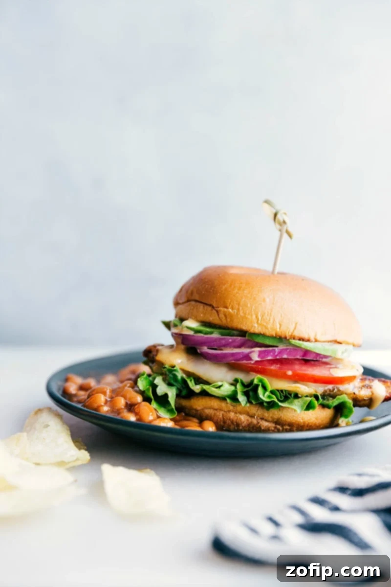 A perfectly assembled chicken burger on a plate, featuring a crispy chicken patty, melted cheese, fresh lettuce, and tomato, served alongside a bowl of baked beans. This delicious and satisfying meal is ready to be enjoyed.