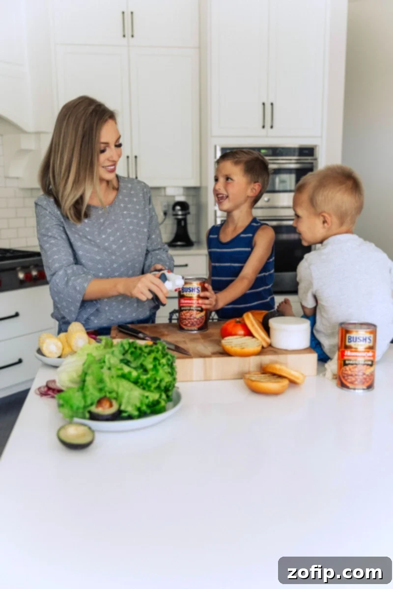 Chelsea and her young sons are depicted in a candid moment, opening a can of baked beans. The scene highlights their involvement in meal preparation, emphasizing the family-friendly nature of the chicken burger recipe and the accompanying side dish.