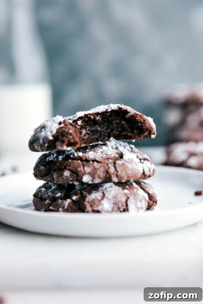 A trio of freshly baked chocolate crinkle cookies, stacked and one split open to beautifully reveal their soft, gooey, and fudgy centers, contrasting with the snowy powdered sugar coating.