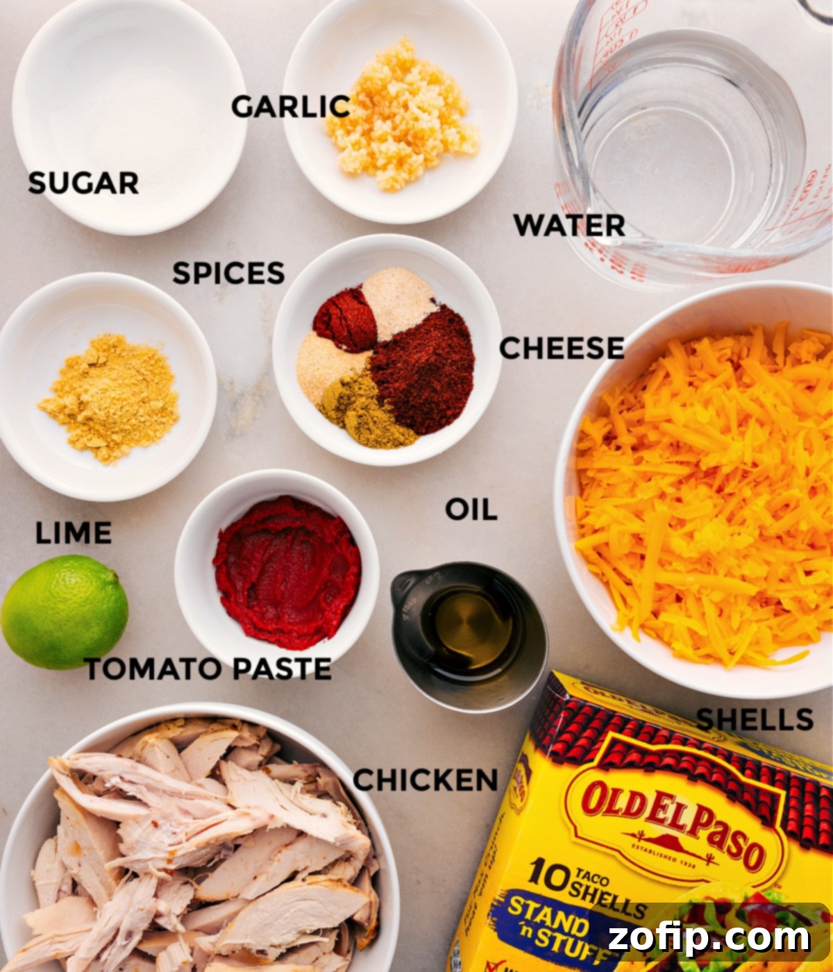 Ingredients In Rotisserie Chicken Tacos Key ingredients for quick Rotisserie Chicken Tacos laid out: shredded chicken, a bowl of water, shredded cheese, crispy taco shells, various taco seasonings, tomato paste, a fresh lime, minced garlic, and a small dish of sugar.
