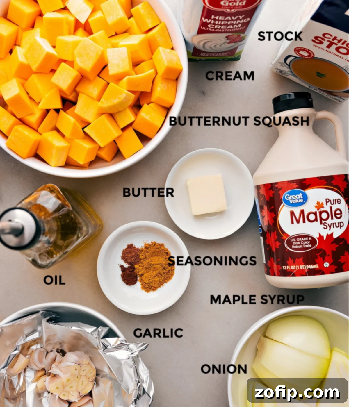 A vibrant display of all fresh ingredients for butternut squash soup, including butternut squash, yellow onion, garlic, spices, broth, cream, maple syrup, and olive oil, neatly arranged on a counter.