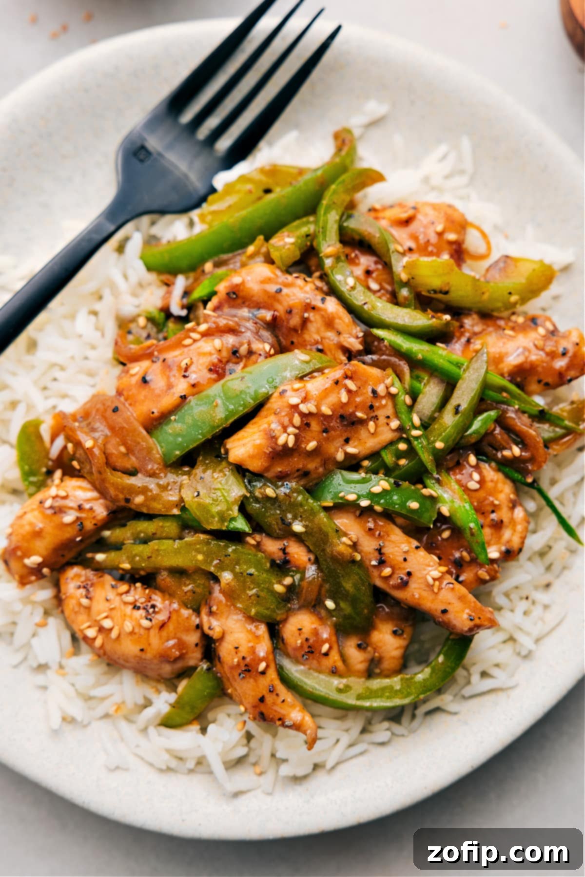 A vibrant plate of Black Pepper Chicken, featuring tender chicken pieces and colorful vegetables coated in a rich, dark sauce, served over white rice and garnished with sesame seeds.