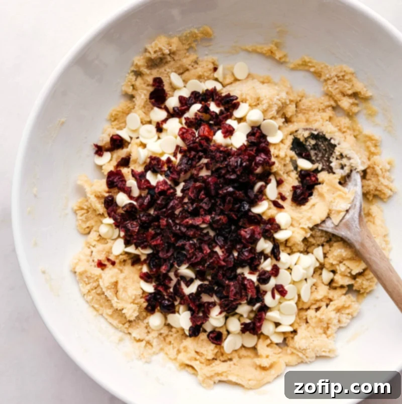 White chocolate chips and cranberries being added to cookie dough in a bowl, enhancing the flavors for a delightful taste combination in these festive cookies.