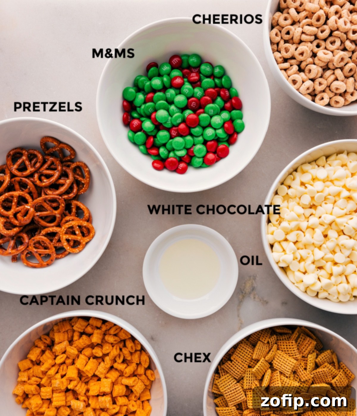 A colorful array of ingredients for Christmas Snack Mix laid out, including various cereals, pretzels, M&M's, white chocolate chips, and a small bottle of oil.