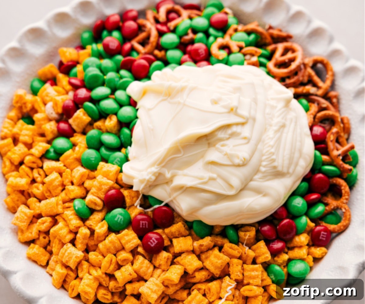 Melted white chocolate being poured over a bowl of mixed cereals, pretzels, and M&M's for the Christmas Snack Mix.