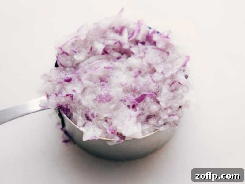 A measuring cup filled with finely grated red onion, showcasing a prepared ingredient for Greek Meatballs.