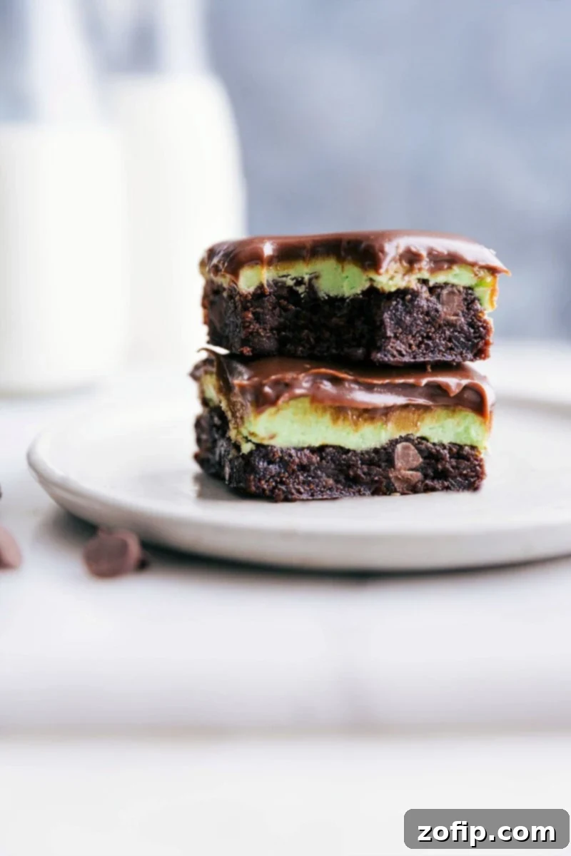 Stacked squares of mint brownies with a bite taken out of the top one, showcasing distinct layers of fudgy brownie, vibrant green mint frosting, and glossy chocolate ganache.