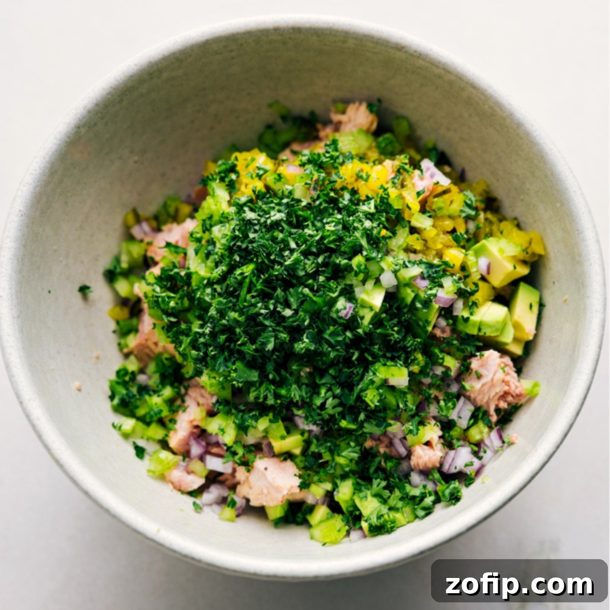 All the fresh ingredients, including flaked tuna, diced celery, red onion, pickles, and parsley, are combined in a large bowl, ready to be mixed with the creamy avocado dressing for this delicious Avocado Tuna Salad.