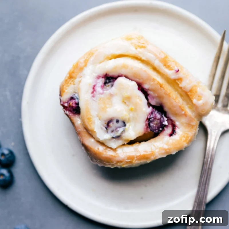 Indulge in a Perfect Lemon Blueberry Sweet Roll A single, perfectly glazed lemon blueberry sweet roll resting on a simple white plate, glistening and ready to be savored with its inviting texture and fresh berries.