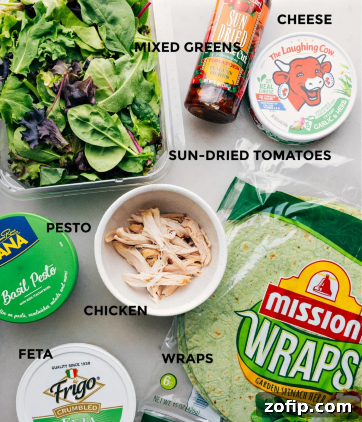 Ingredients In Mediterranean Wrap Recipe All the fresh ingredients for a Mediterranean Wrap, including pesto, shredded chicken, mixed greens, sun-dried tomatoes, feta, and tortillas, laid out on a clean countertop.