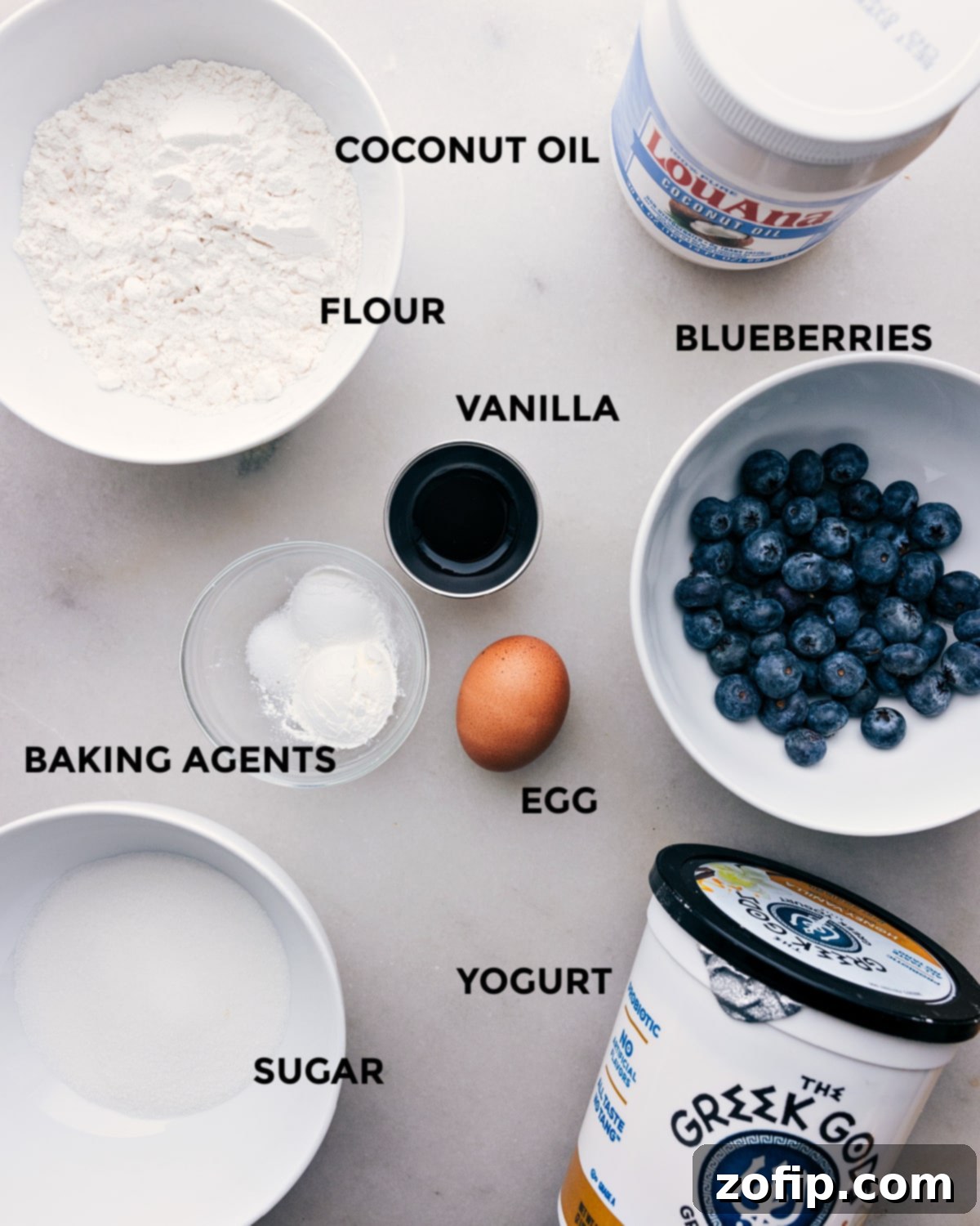 Wholesome Ingredients for Healthy Blueberry Muffins All the wholesome ingredients, including flour, cornstarch, baking powder, salt, blueberries, egg, Greek yogurt, vanilla extract, coconut oil, and light brown sugar, meticulously laid out on a counter for healthy blueberry muffins.