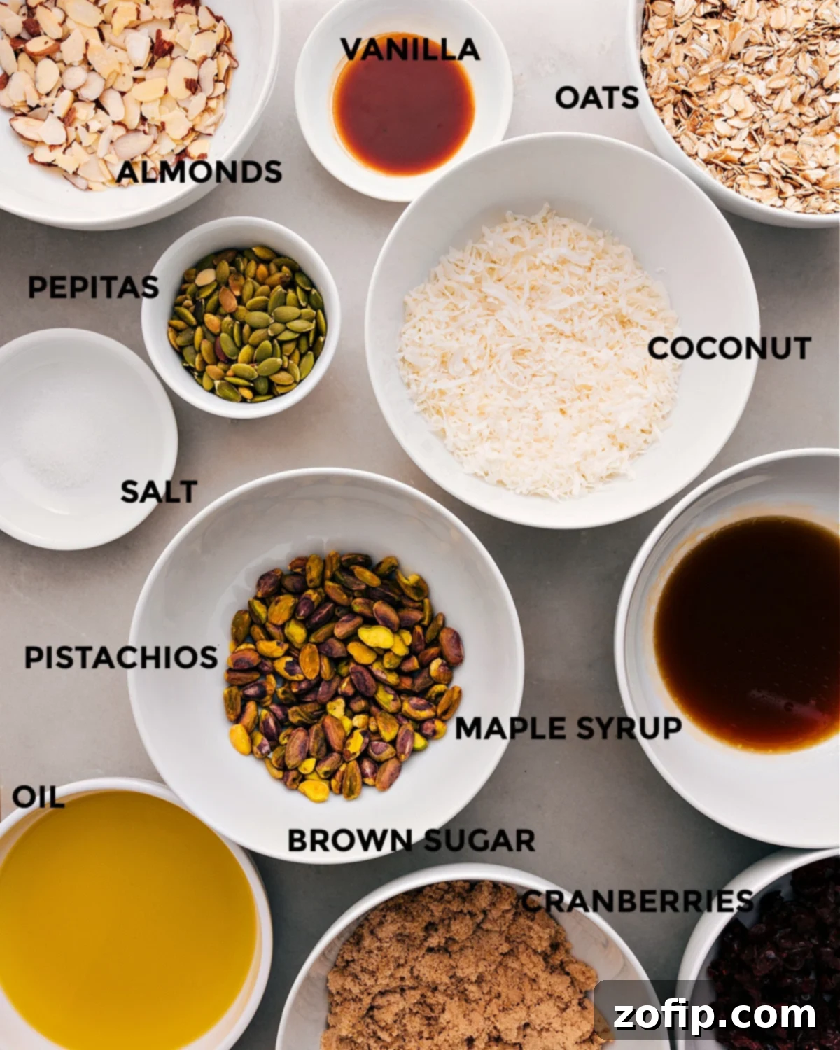 All the fresh and wholesome ingredients laid out for easy assembly: coconut flakes, old-fashioned oats, vanilla extract, sliced almonds, pepitas, pure maple syrup, pistachios, sea salt, brown sugar, dried cranberries, and high-quality olive oil.