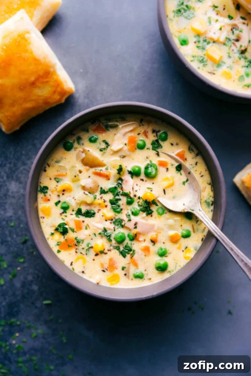 A steaming bowl of Chicken Pot Pie Soup, brimming with shredded chicken, golden potatoes, and colorful vegetables, garnished with fresh herbs and a crisp, golden puff pastry square resting on the rim.