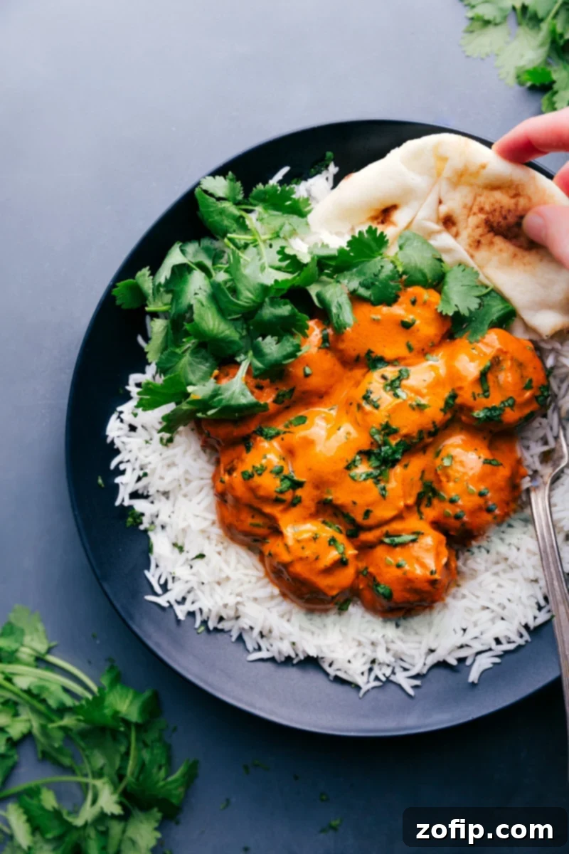 Plate of perfectly cooked Butter Chicken Meatballs served on a bed of fluffy basmati rice, generously garnished with fresh cilantro, accompanied by warm, pillowy naan bread on the side. The rich, orange-hued sauce is visibly creamy and inviting.