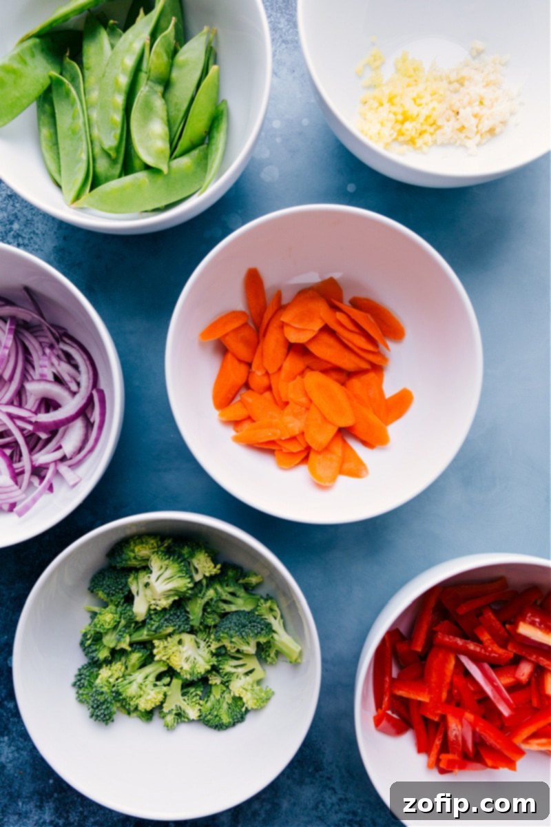Various fresh ingredients for General Tso Chickpeas laid out, including chopped carrots, broccoli florets, snow peas, red bell pepper, ginger, garlic, and a bowl of chickpeas.