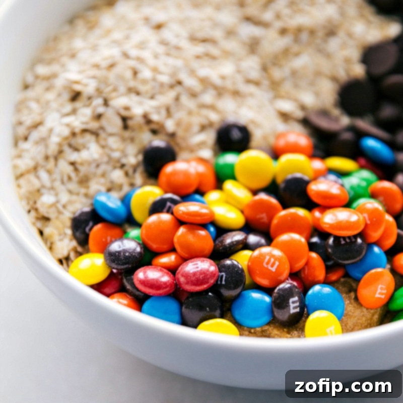 Oats, chocolate chips, and M&M's being added to the bowl with the cookie dough, ready for final mixing to create the perfect monster cookies.