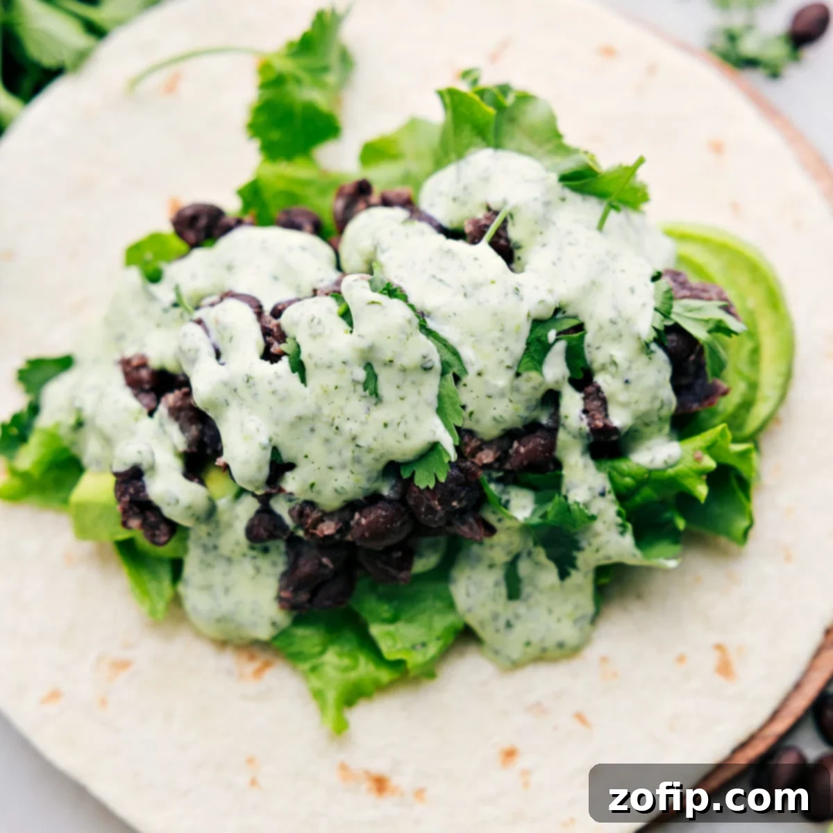 An appetizing black bean wrap, beautifully opened to reveal its vibrant layers of creamy avocado, crisp lettuce, hearty black beans, and the signature flavorful cilantro-lime sauce, ready to be enjoyed.