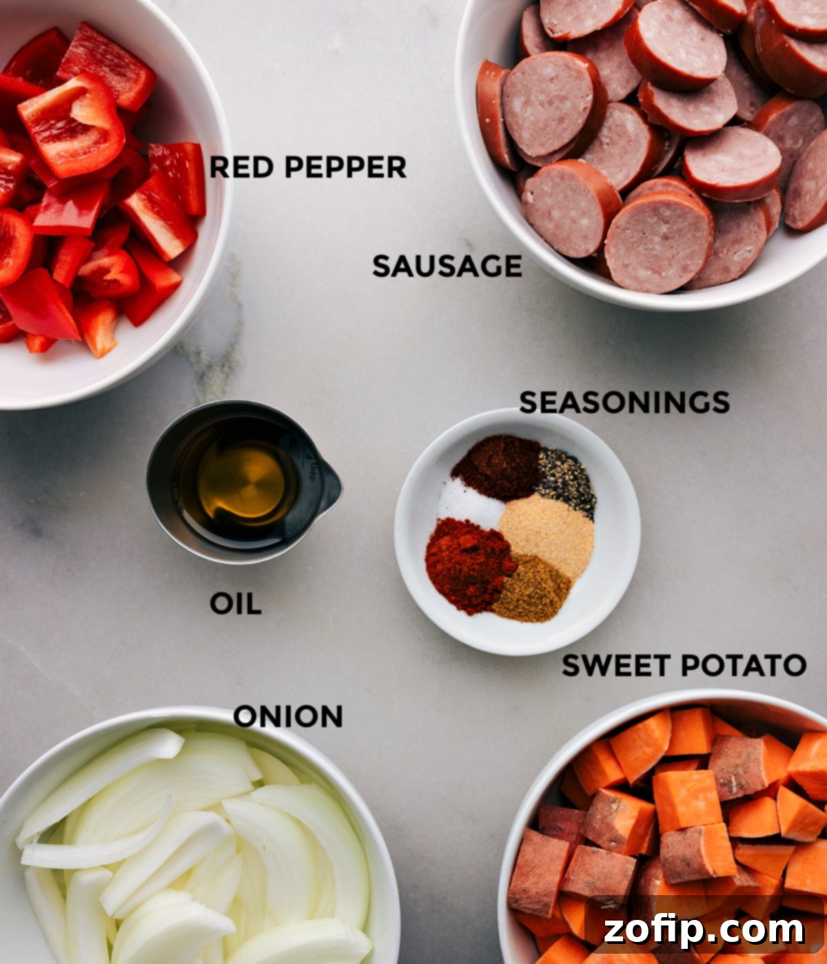 An organized tray of raw ingredients for a delicious one-pan meal, featuring thinly sliced yellow onion, cubed sweet potatoes, vibrant red bell pepper, olive oil, and a selection of spices like chili powder, cumin, and paprika, alongside sliced smoked sausage, ready for roasting.