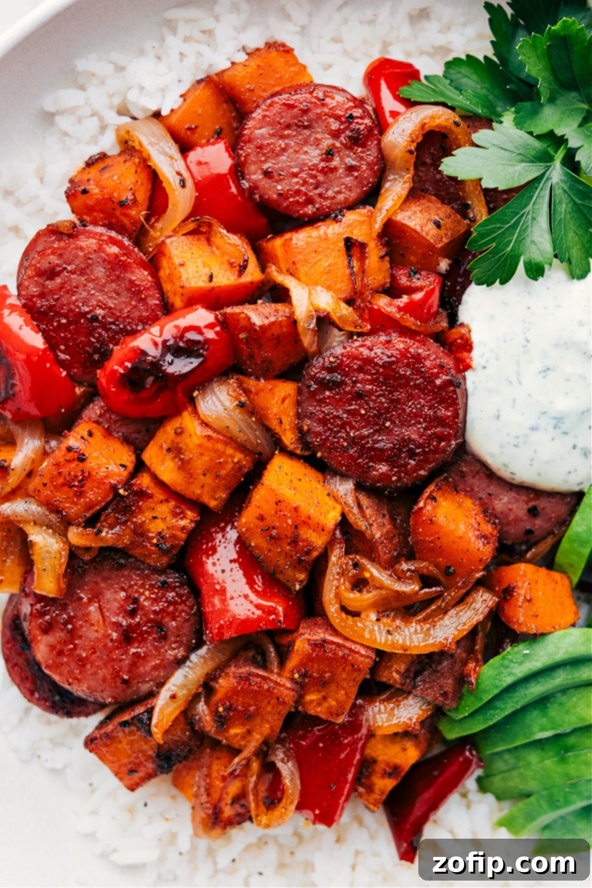 A hearty portion of roasted sweet potato and sausage, beautifully arranged over a bed of fluffy rice, generously drizzled with a creamy green sauce and ready to be served on a white plate.