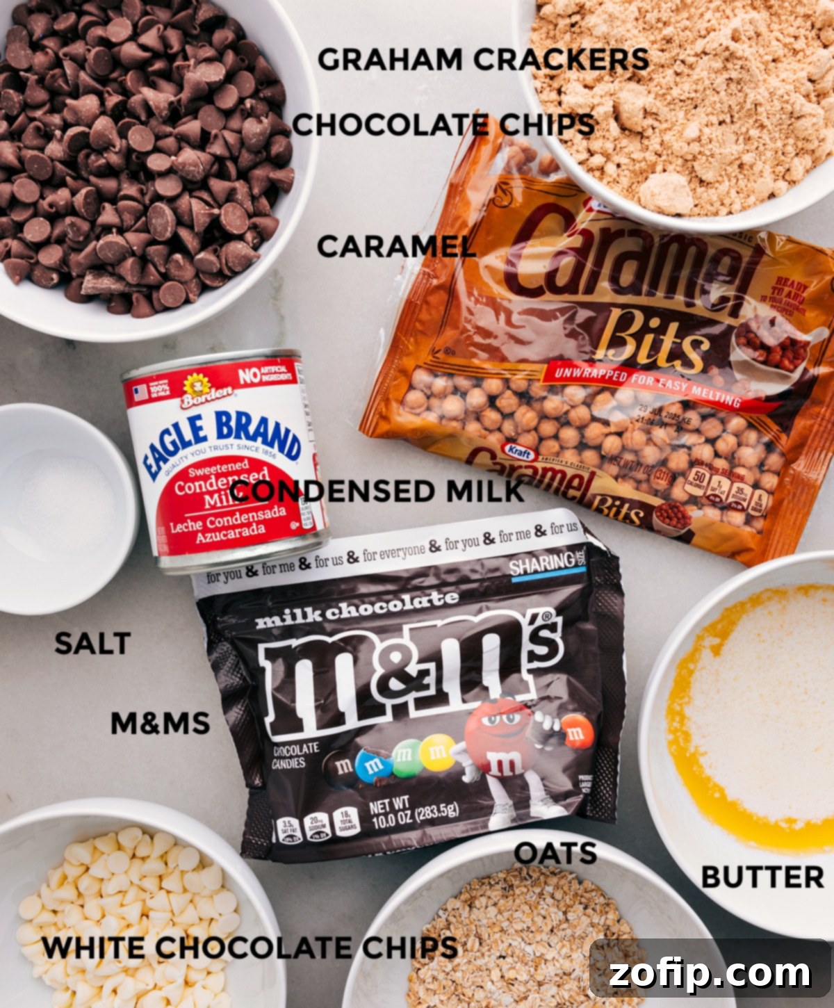 A flat lay showing all the essential ingredients for Magic Cookie Bars: graham crackers, quick oats, chocolate chips, white chocolate chips, caramel bits, M&Ms, sweetened condensed milk, butter, and a pinch of salt.