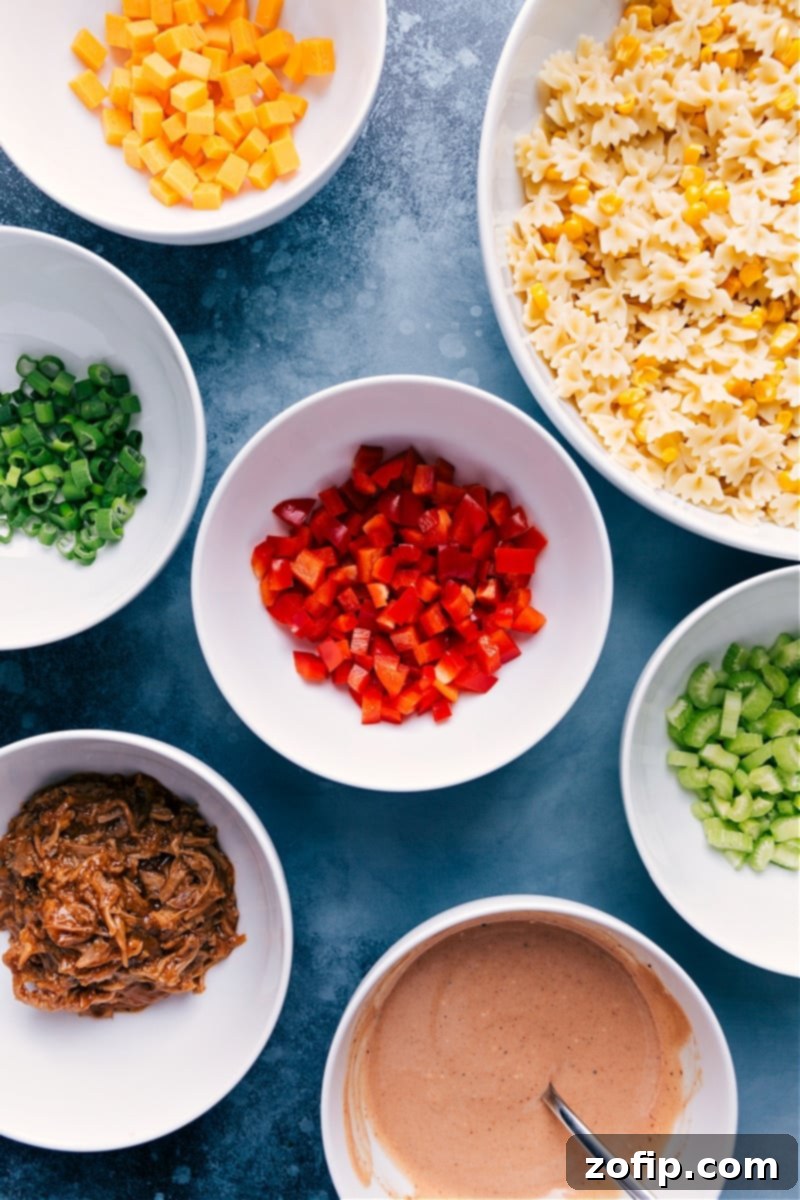 A beautiful spread of fresh ingredients for BBQ Pasta Salad, including perfectly cooked pasta, vibrant bell peppers, crisp celery, green onions, cubed sharp cheddar, succulent pulled pork, and a bowl of the creamy BBQ dressing.