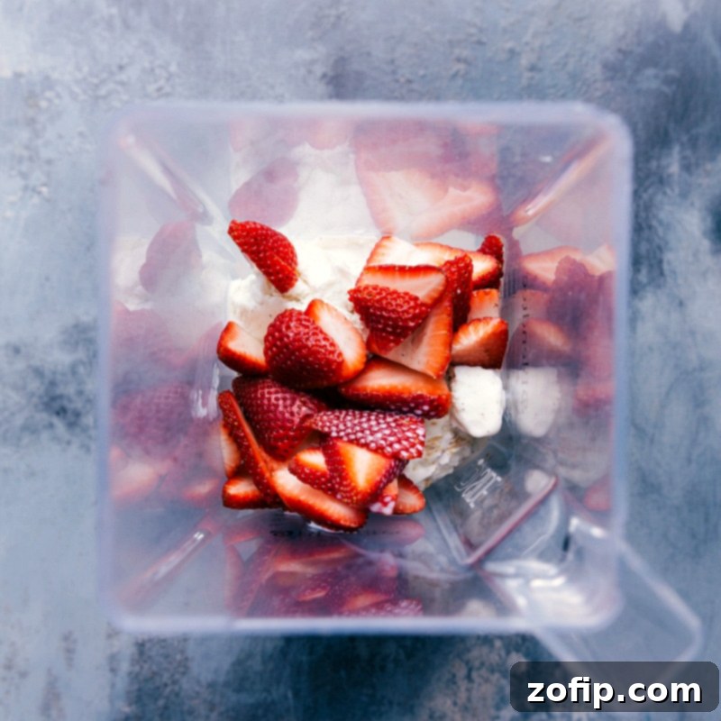 All four essential ingredients for a perfect strawberry milkshake – fresh, hulled strawberries, a splash of vanilla extract, scoops of creamy vanilla bean ice cream, and whole milk – being carefully added into a high-powered blender jar, ready for mixing.