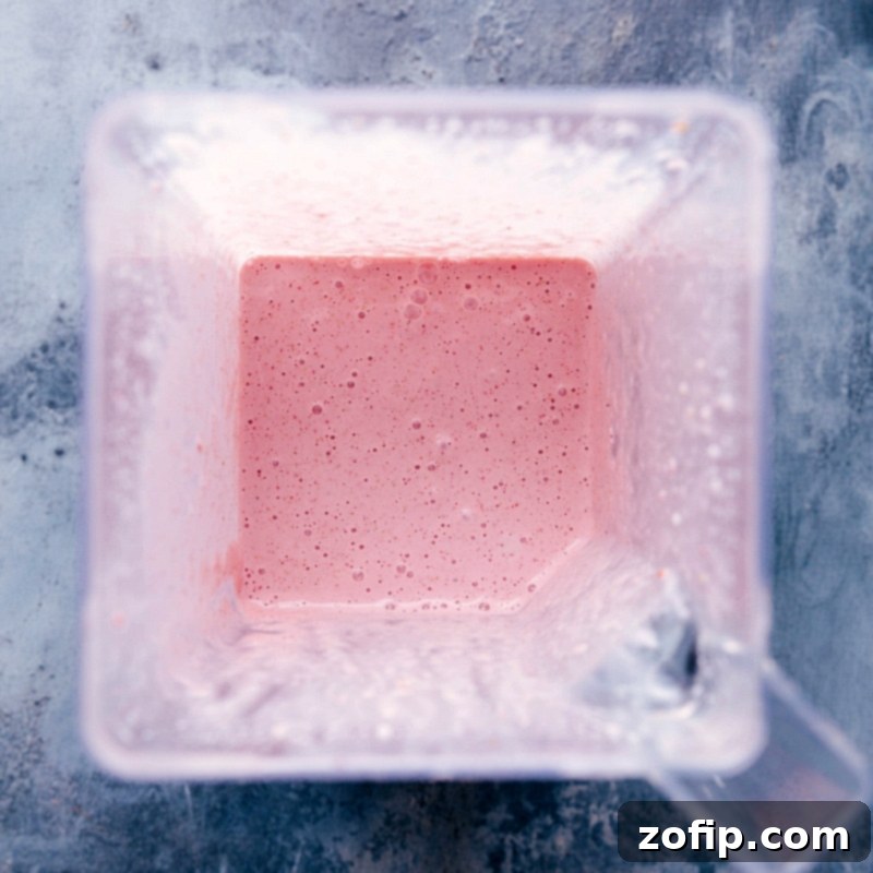 The perfectly blended strawberry milkshake, smooth, frothy, and a beautiful pink hue, rests serenely in the blender jar, signaling it's ready to be poured into a chilled glass and enjoyed.